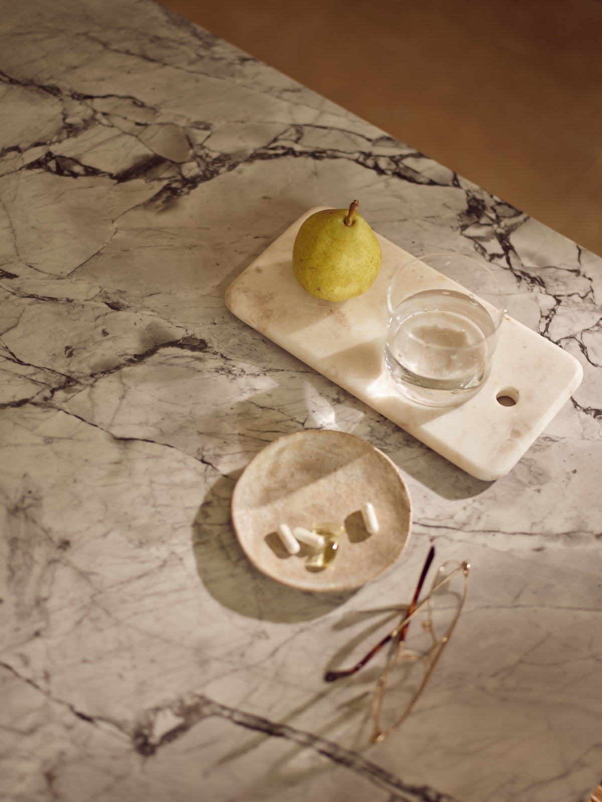Marble surface with a small dish with different supplement capsules on it and next this is a  pear and glass of water.