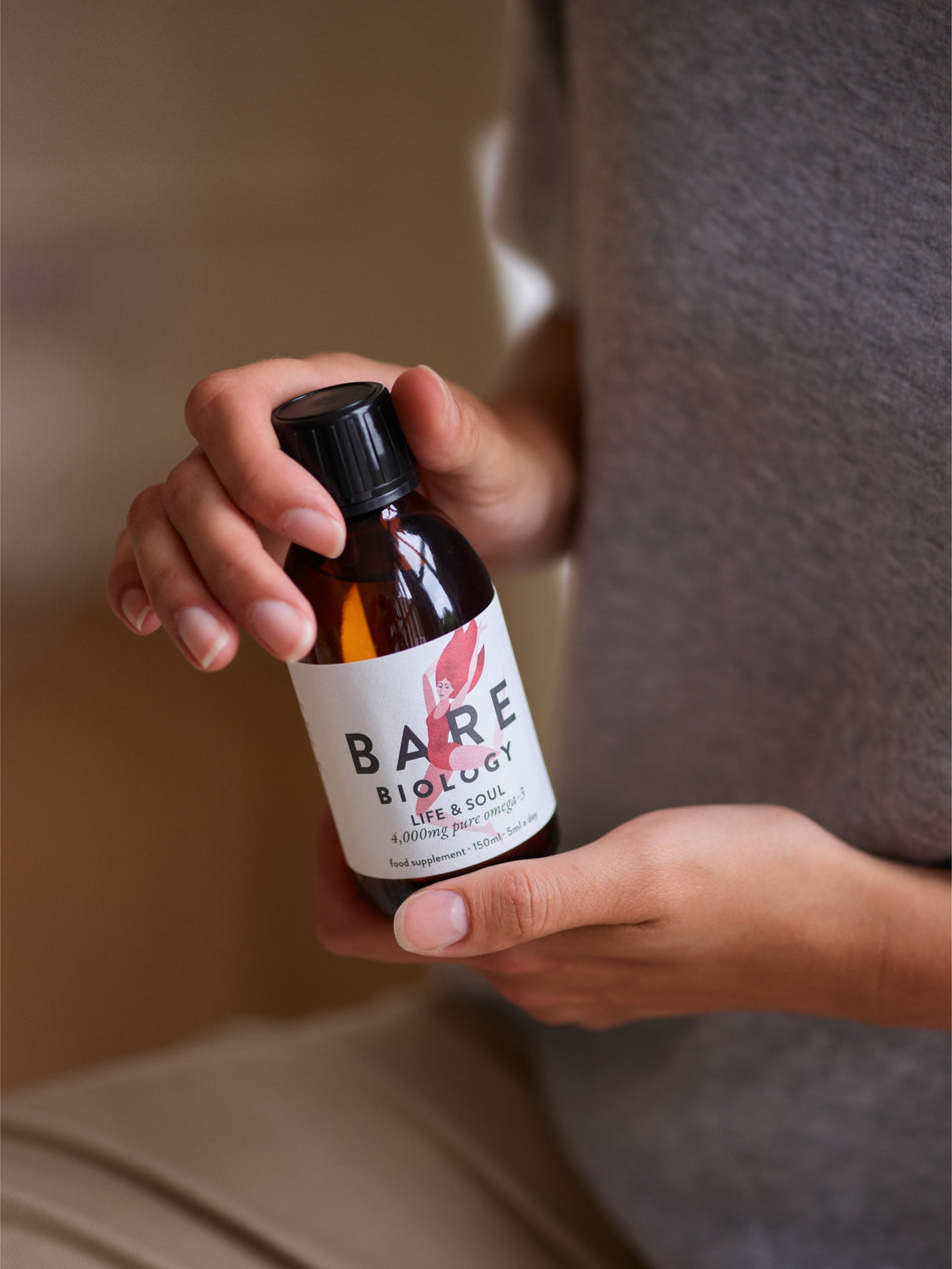 Hand holding a bottle of life and soul omega-3 fish oil labeled with 'BARE BIOLOGY' against a neutral background