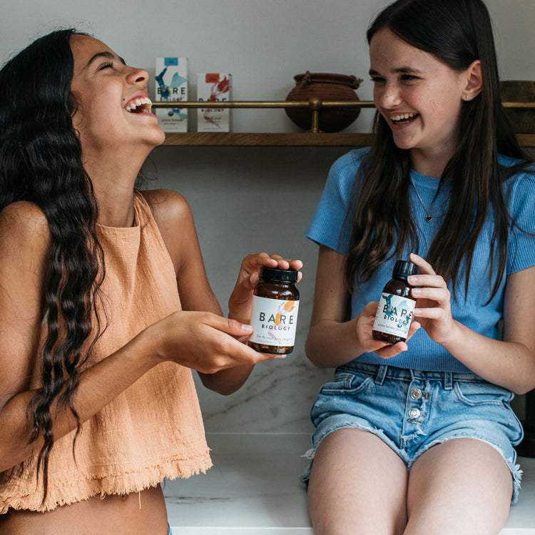 Two teens laughing looking at each other and holding bottles of Bare Biology Omega-3 products.