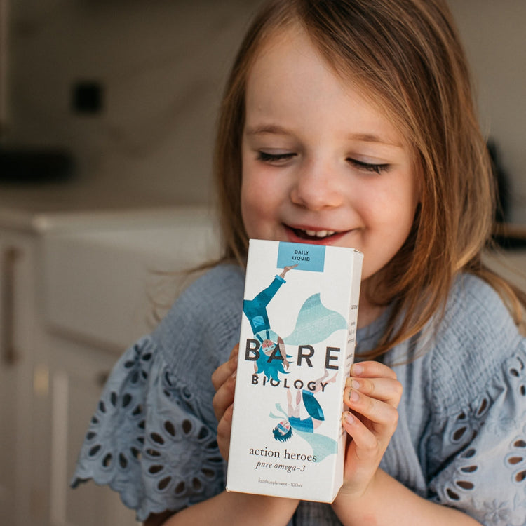 Little girl holding a Bare Biology Action Heroes liquid Omega-3 supplement box with blue illustrations on the front