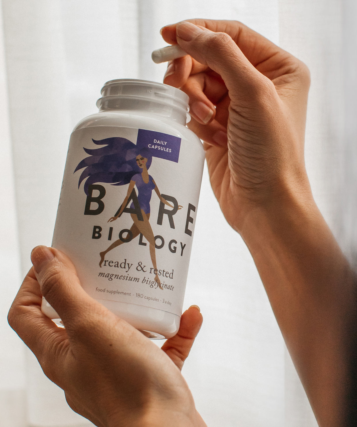 Close up of a woman's hands holding a pot of Bare Biology magnesium in one hand and pulling out a capsule with the other.