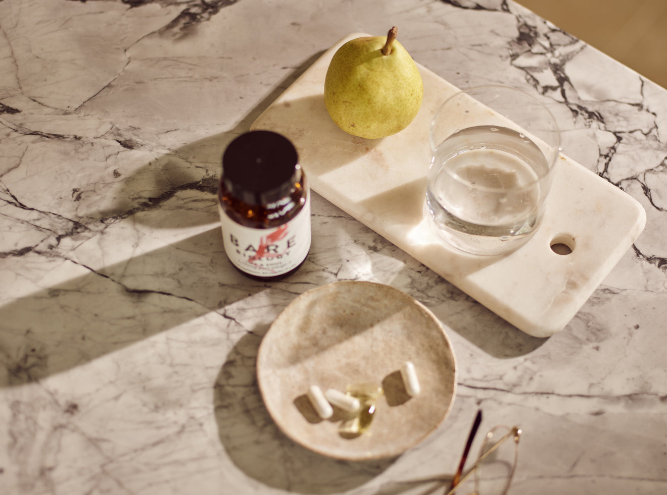 Bottle of Bare Biology Daily omega-3 capsules on a marble surface with a pear and glass of water.