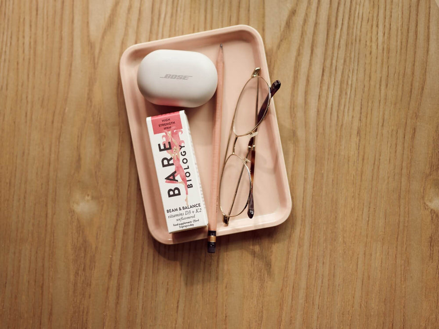Image of pink tray with bose wireless earphones, pencil, reading glasses and Bare Biology Beam and Balance spray placed on top