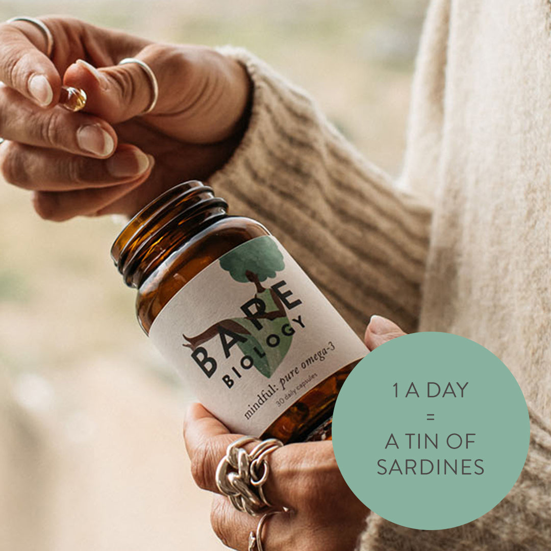 A lady with a beige jumper and silver rings taking a capsule out of a Mindful DHA omega-3 jar. Dose advise listed in a circle: 1 a day = a tin of sardines.
