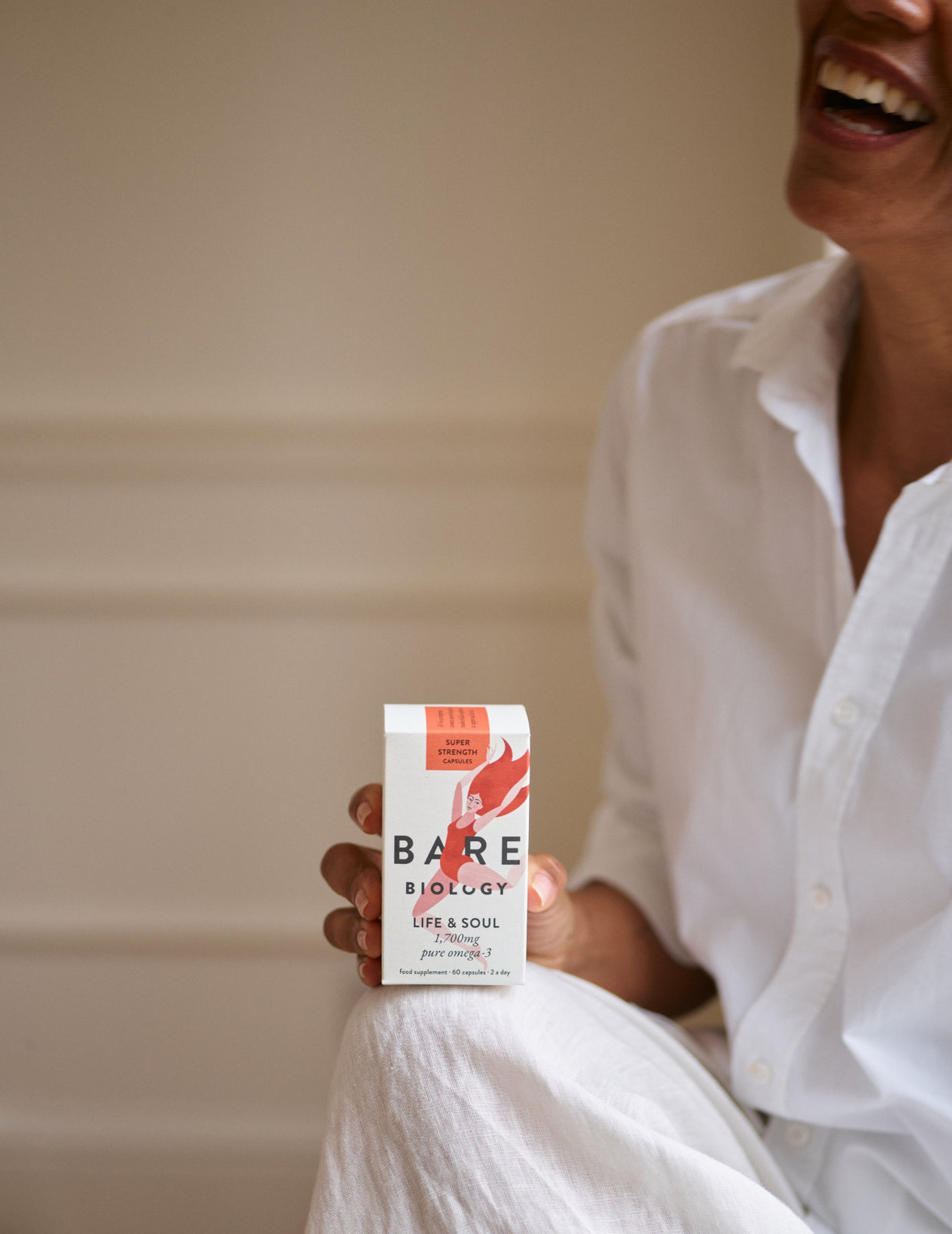 Cropped lady wearing white linen clothing smiling and holding a box of Life & Soul Daily capsules on her knee