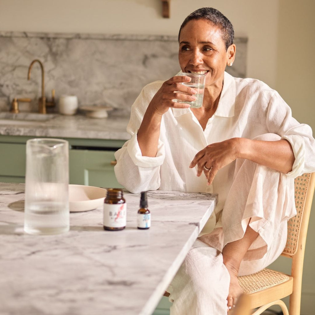 Woman sitting at a kitchen table with bare biology products and water glasses and bottles, holding a glass of water.
