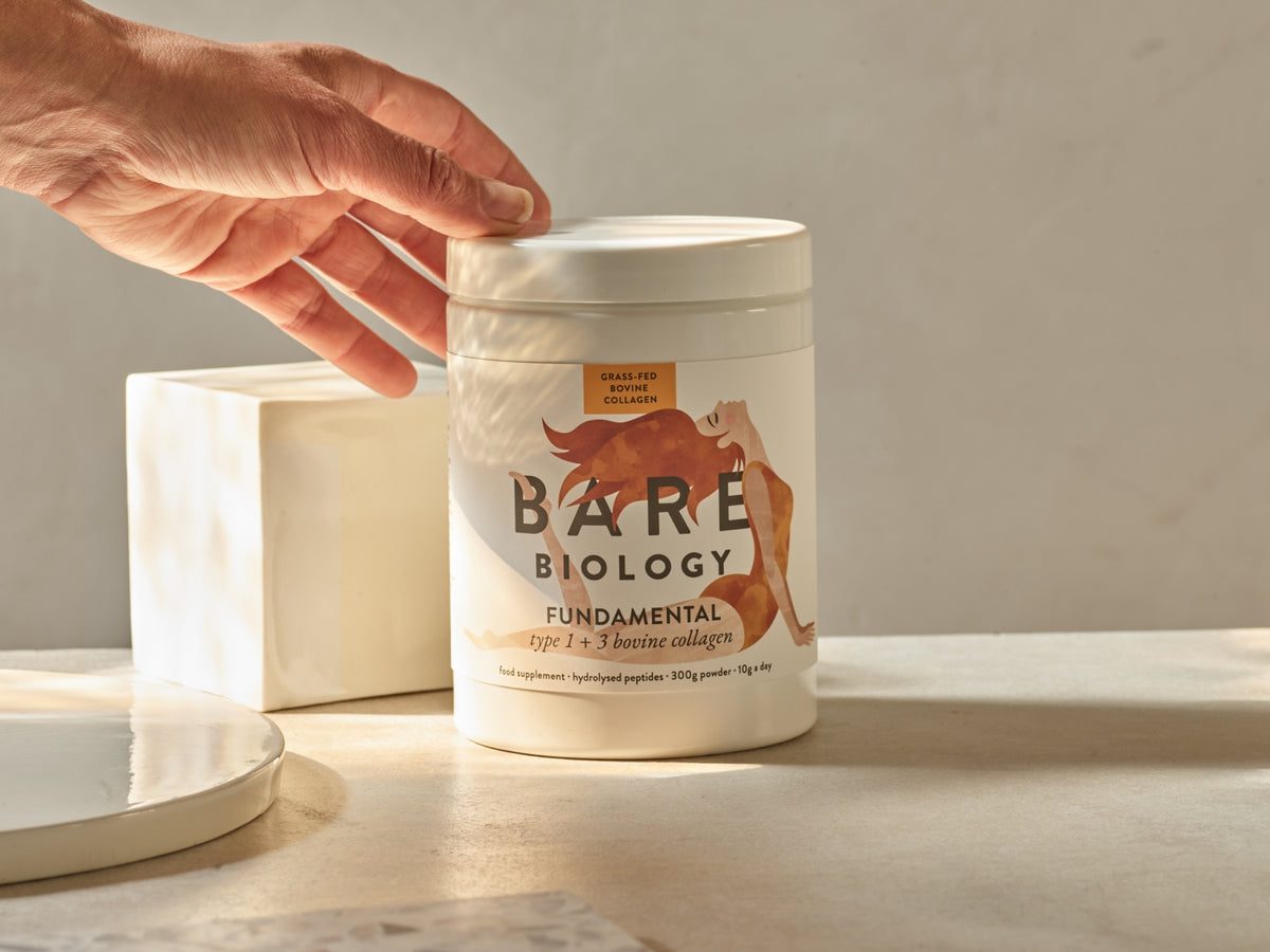 a woman's hand going to grab a white tub labelled with Bare Biology Fundamental Bovine collagen. The collagen is on a beige stone table and there are light shadows coming in from the left.