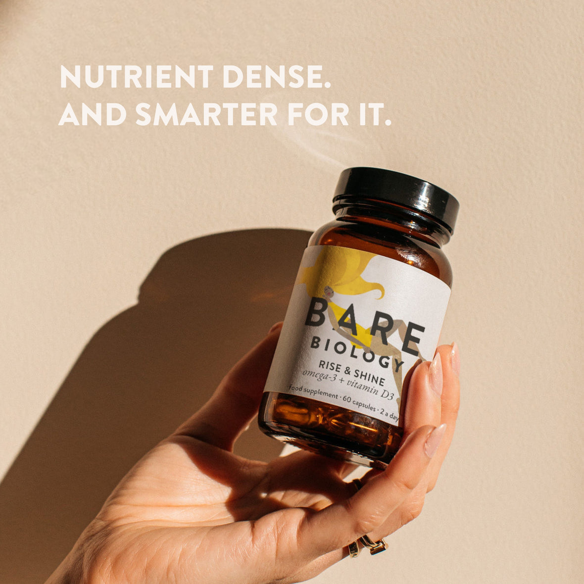 Hand holding a bottle of Bare Biology supplements with a beige background and text 'NUTRIENT DENSE. AND SMARTER FOR IT.'