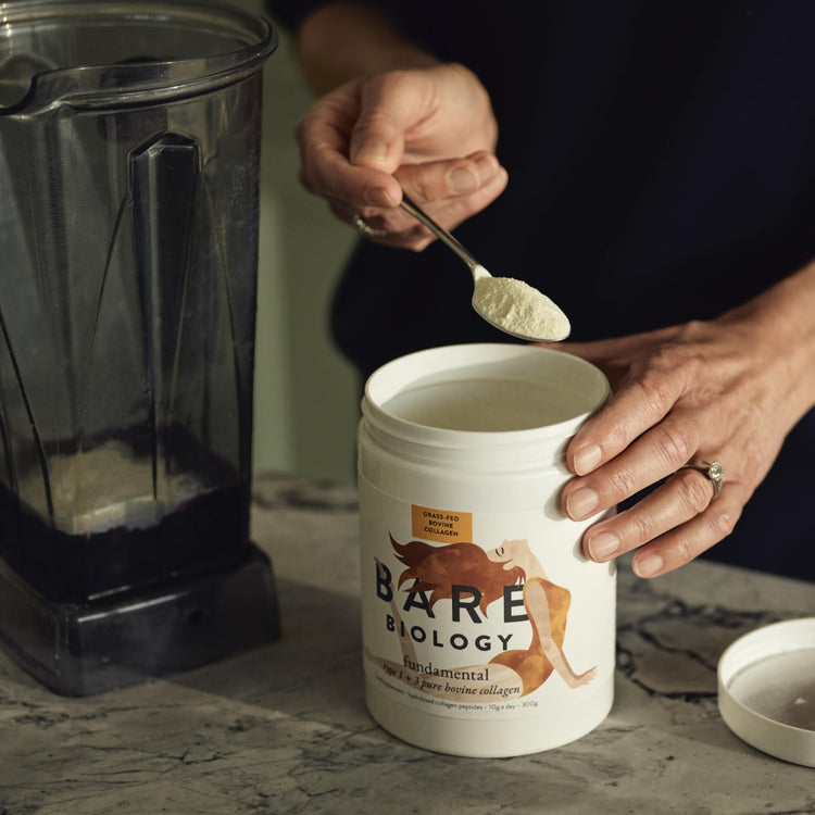 Lady scooping Bare Biology bovine collagen powder with a spoon from white product tub. There is a smoothie blender container next to her.