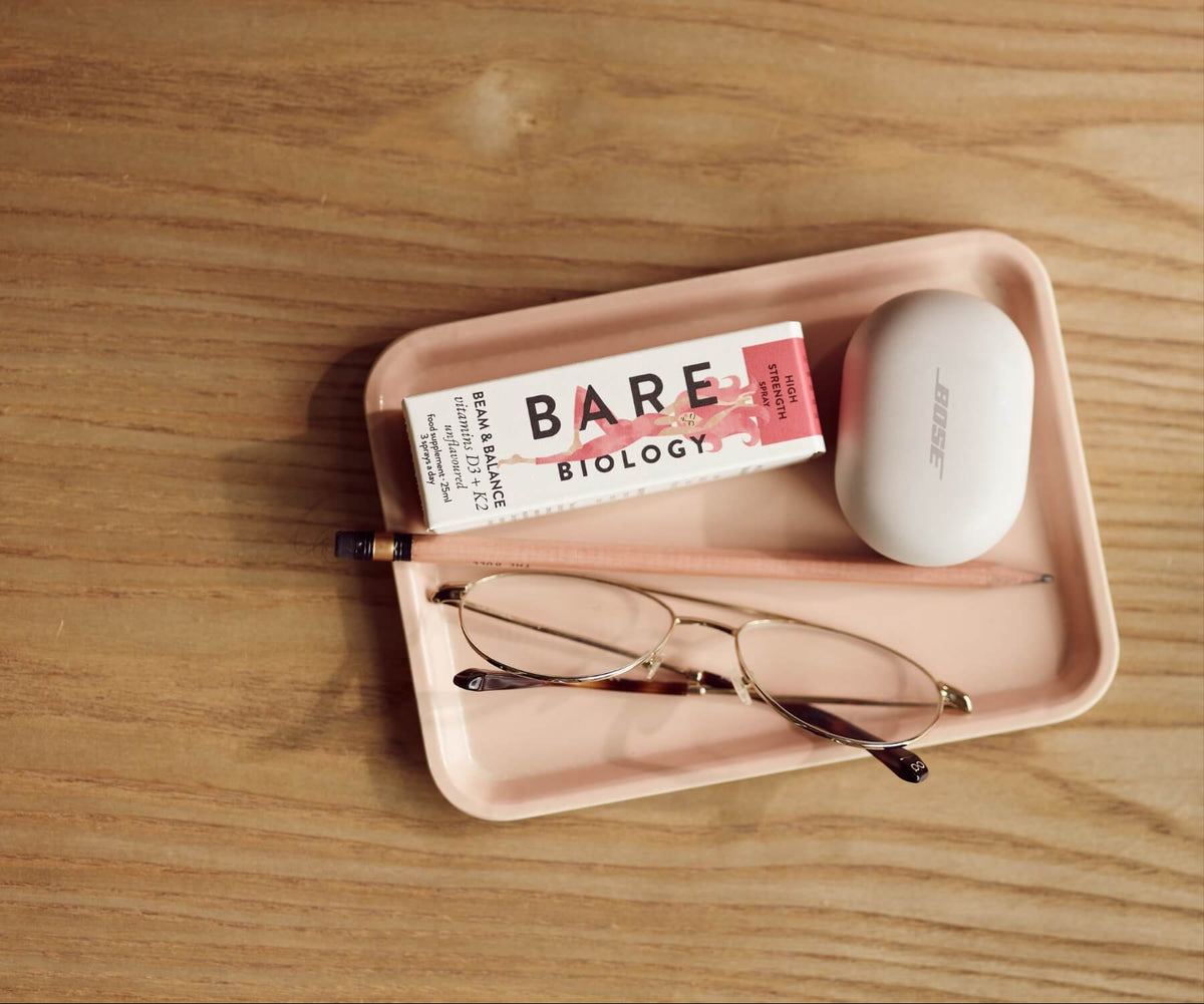 Pink tray with a carton of Bare Biology Vitamin D3+K2, glasses, and a white bar of soap on a wooden surface.