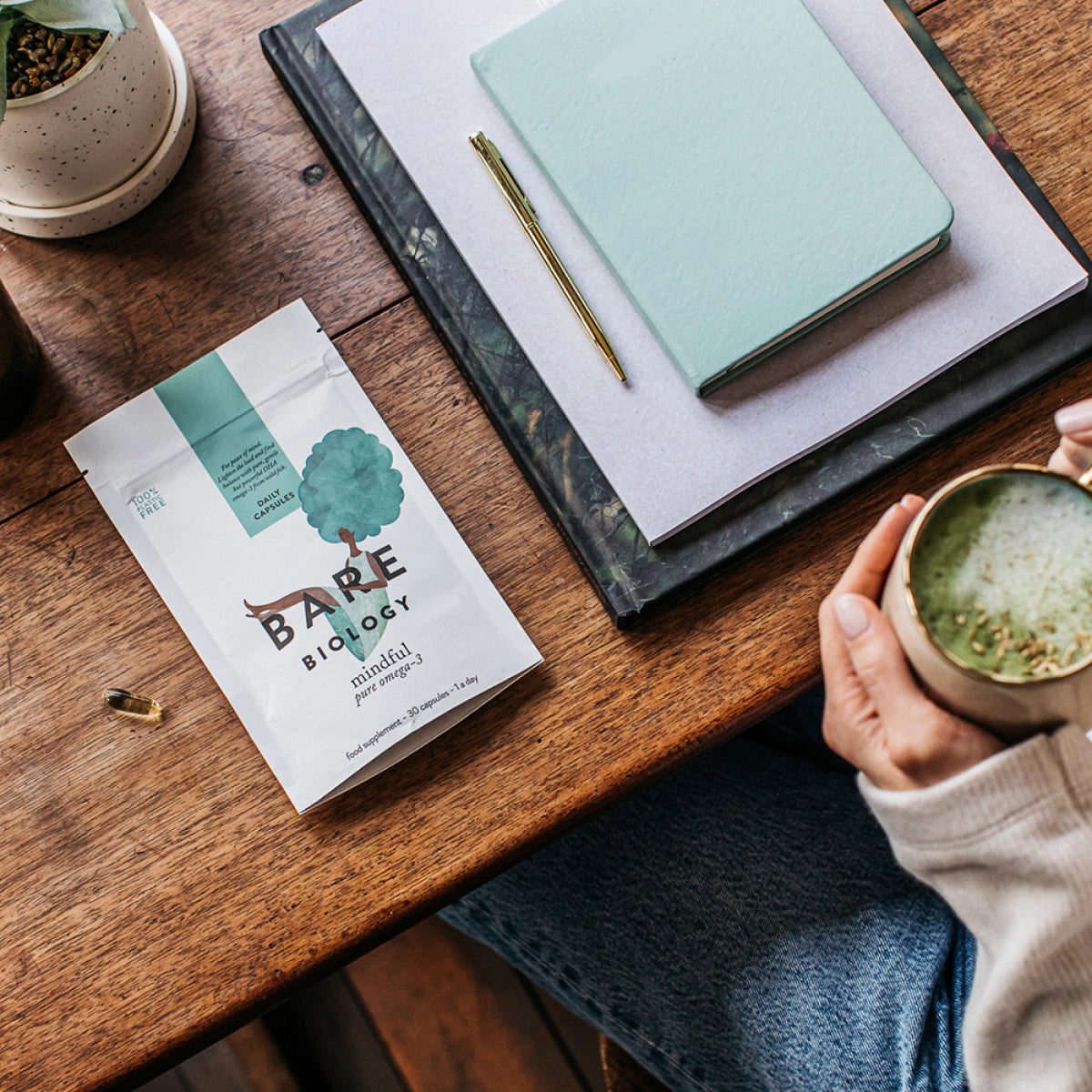woman at a desk holding a matcha tea with a pouch of bare biology mindful omega capsules and notebooks on the table