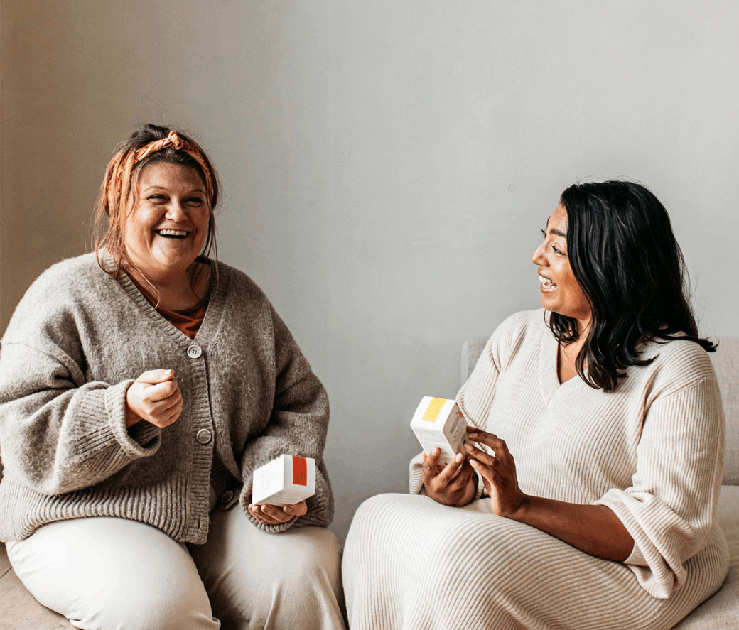 Two woman smiling facing forward and to each other holding bare biology products on a neutral coloured background