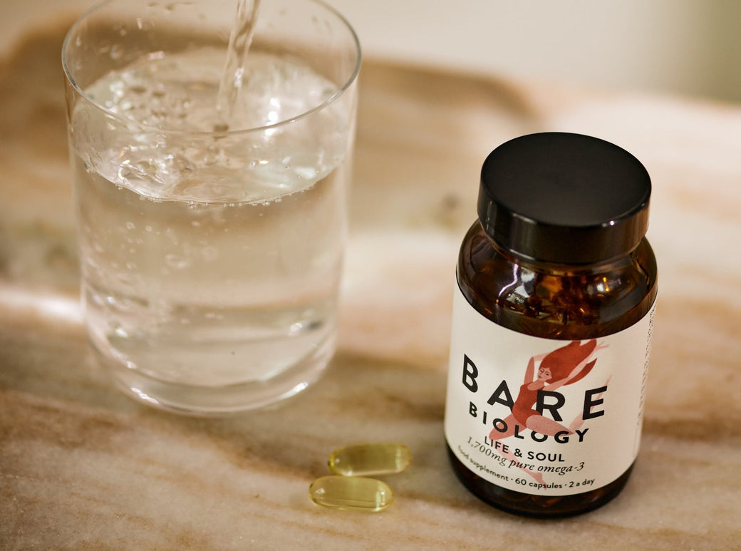 Jar of Bare Biology supplements with a glass of water and two supplement capsules displayed on a marble surface