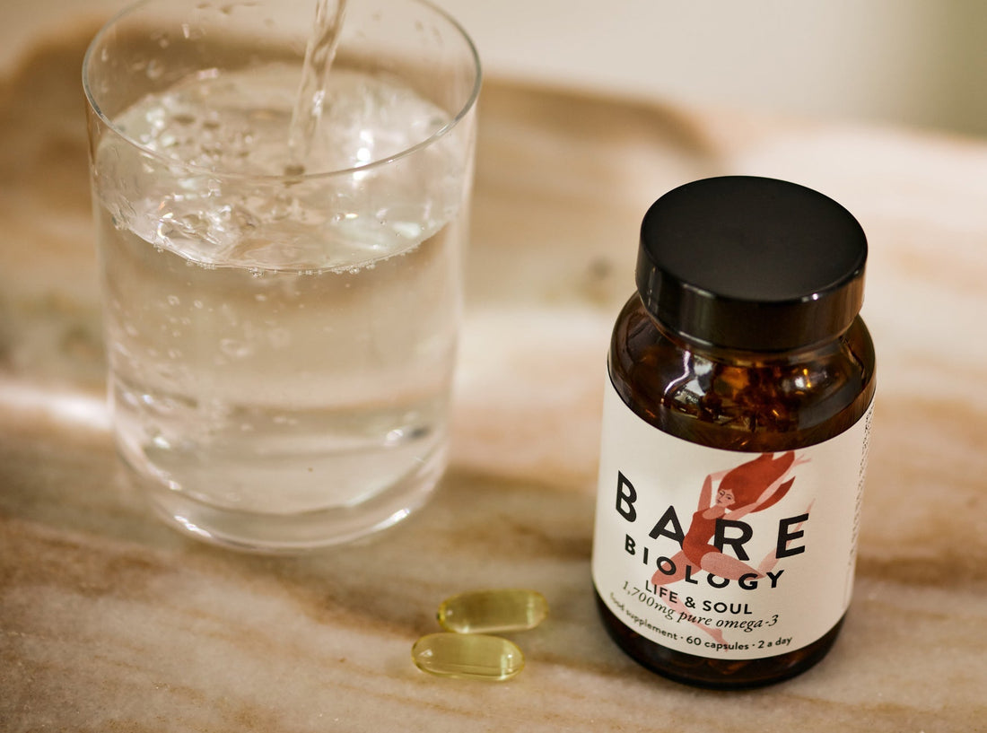 Jar of Bare Biology supplements with a glass of water and two supplement capsules displayed on a marble surface