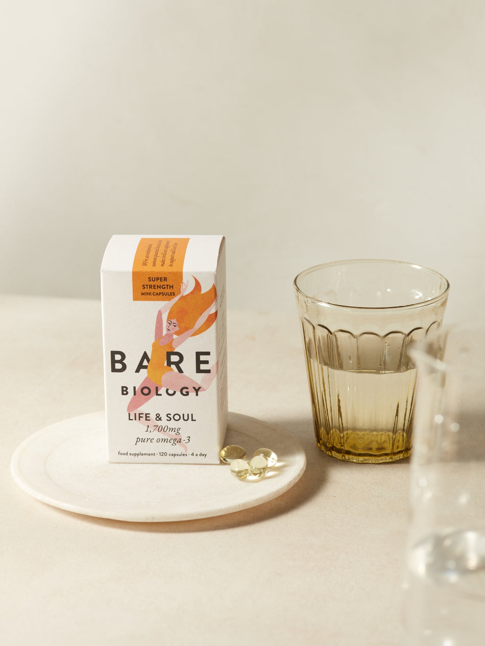 Bare Biology omega-3 mini supplement capsules box with a glass of water on a neutral background, and 4 capsules by the side of the box