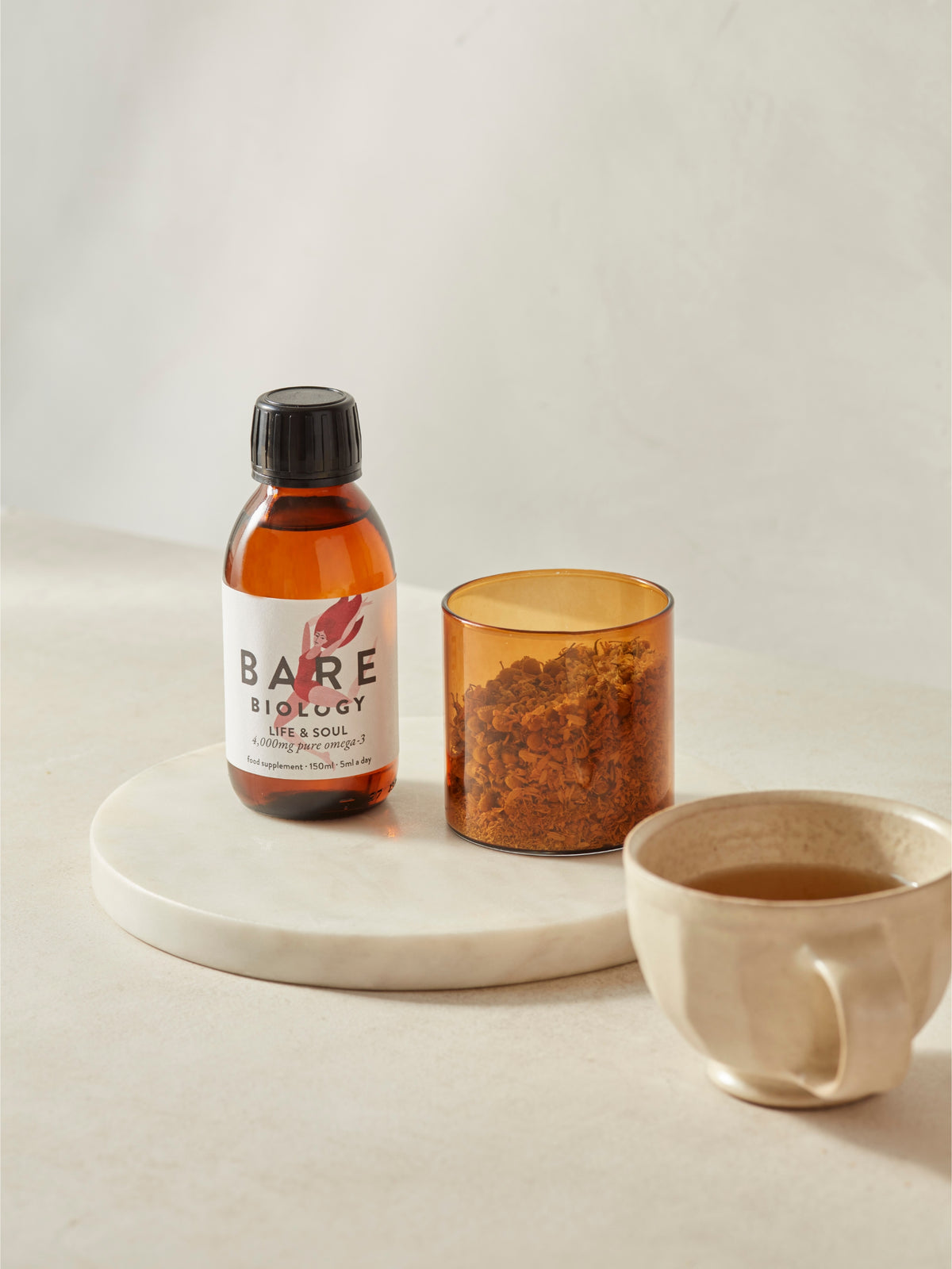 Bare Biology fish oil liquid glass bottle on a marble tray with a candle and mug on a light background