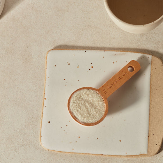 Wooden 10g bare biology measuring scoop with collagen powder on a ceramic plate, with a cup of tea in the background.