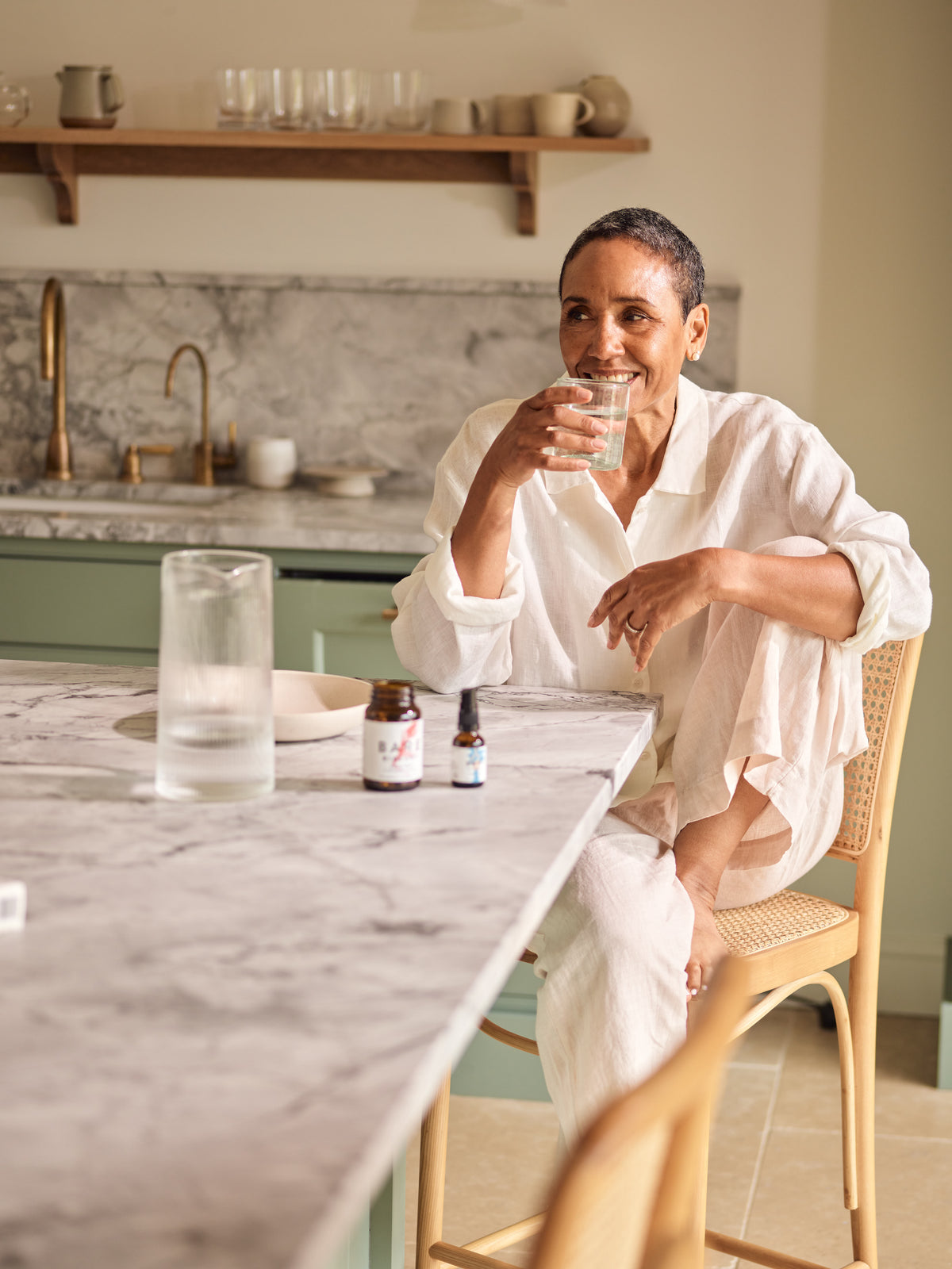 Woman sitting at a kitchen counter drinking water with her supplements with a neutral background in a rustic and neutral looking kitchen.