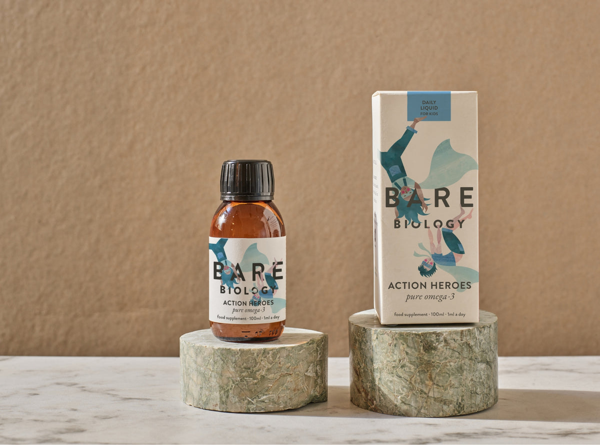 Bare Biology Action Heroes Liquid for children in a bottle and next to it in its's packaging on a neutral background displayed on ceramic style circular stands