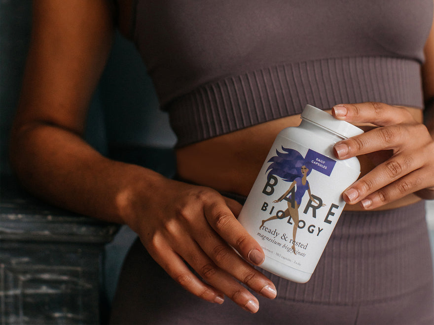 close up of a woman in gym leggings and a sports bra. The camera is focused in on a tub of Bare Biology magnesium capsules which she's holding at waist height.