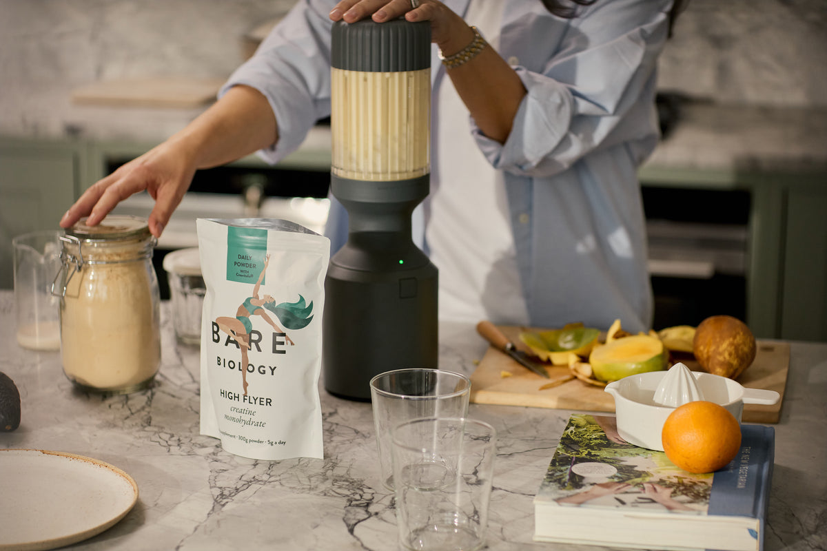 Person using a blender with smoothie ingredients on a kitchen counter, including a jar of cream and a package labeled 'Bare Biology'.