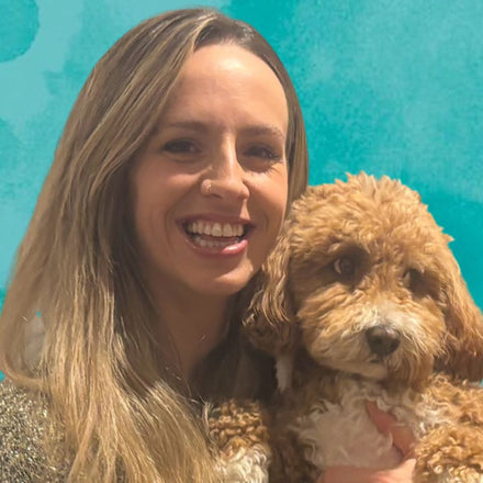 Becky smiling with long blonde hair holding a small curly-haired brown dog Ziggy, against a bright turquoise background.