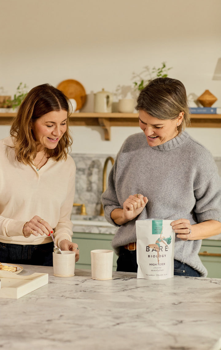 Two woman interacting with one another whilst in a kitchen setting, whilst one stirs the cups of tea the other has opened a pouch of Bare Biology's creatine.