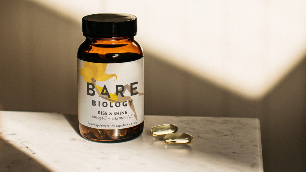 Bottle of Bare Biology Rise & Shine capsules next to two capsules on a marble surface with sunlight casting shadows.
