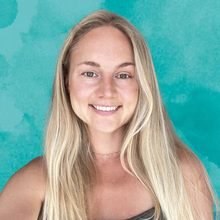 A smiling blonde lady with green eyes wearing a delicate gold necklace, set against a textured teal watercolor background.