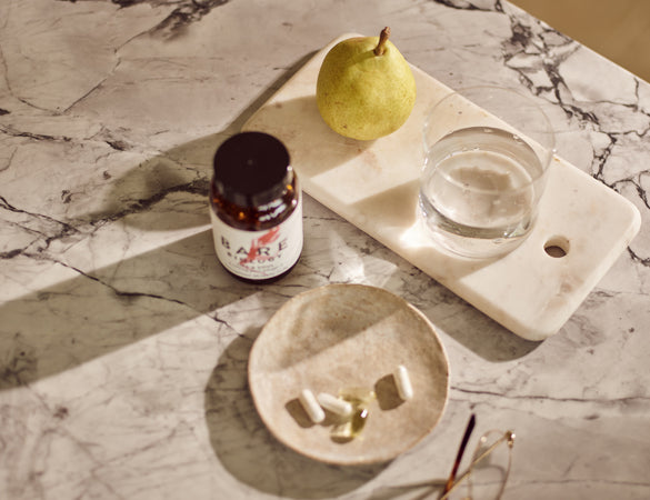 Bottle of Bare Biology Daily omega-3 capsules on a marble surface with a pear and glass of water.