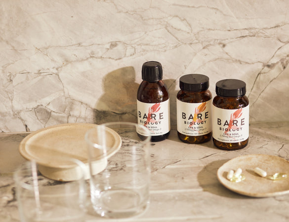 Bare Biology Life & Soul range; bottles of capsules and liquid on a marble top with marble background and a ceramic plate of capsules and glass in front of them.  