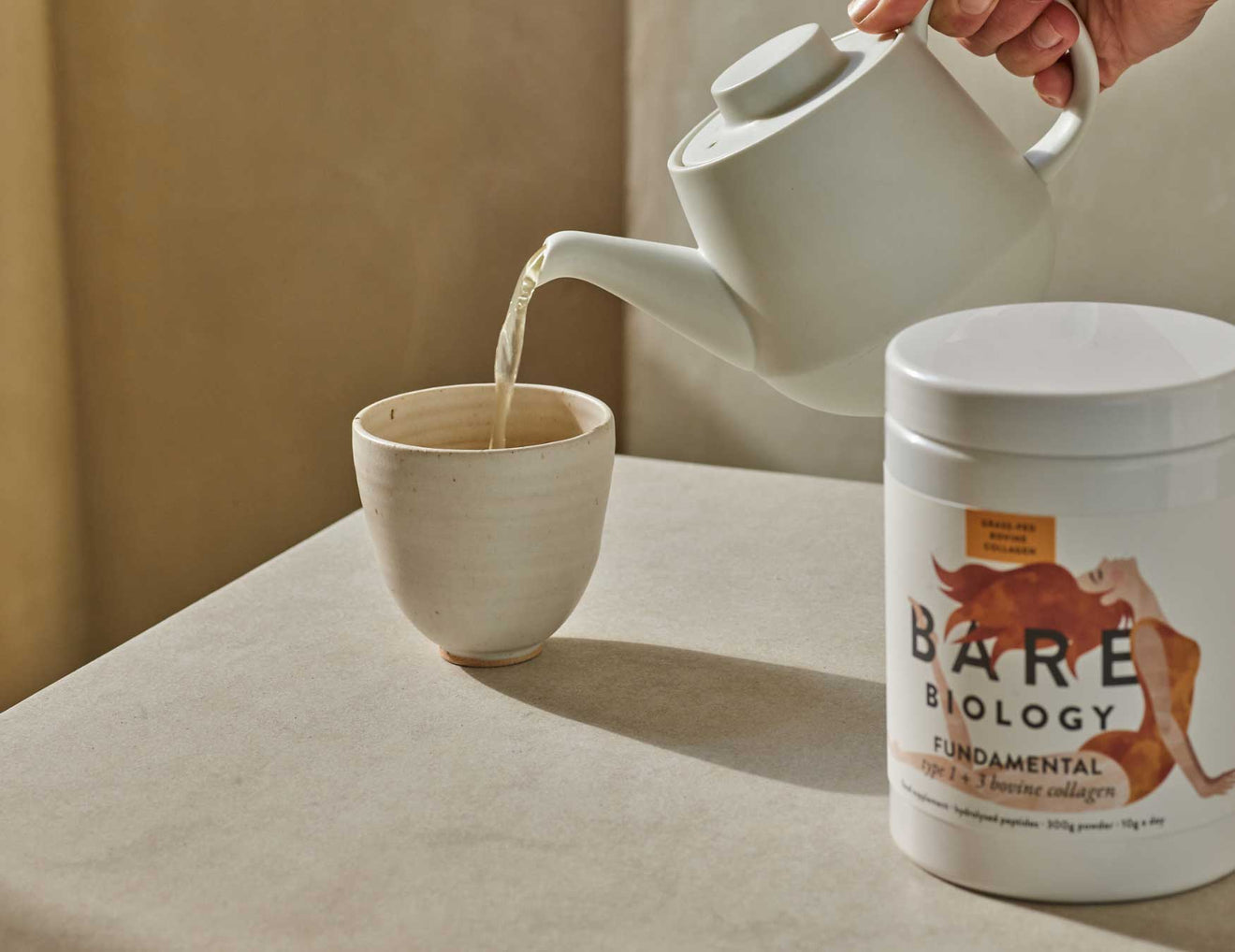 Person pouring tea from a white teapot into a ceramic cup on a beige surface, with a container of Bare Biology powder in the foreground.