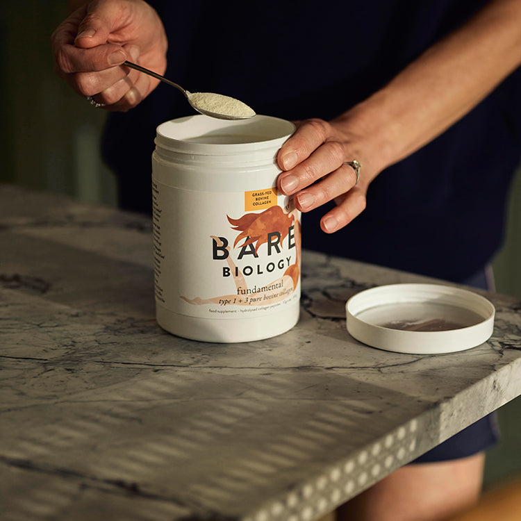 Woman scooping Fundamental Bovine Collagen powder from white tub with s spoon.The tub has Bare Biology branded label.