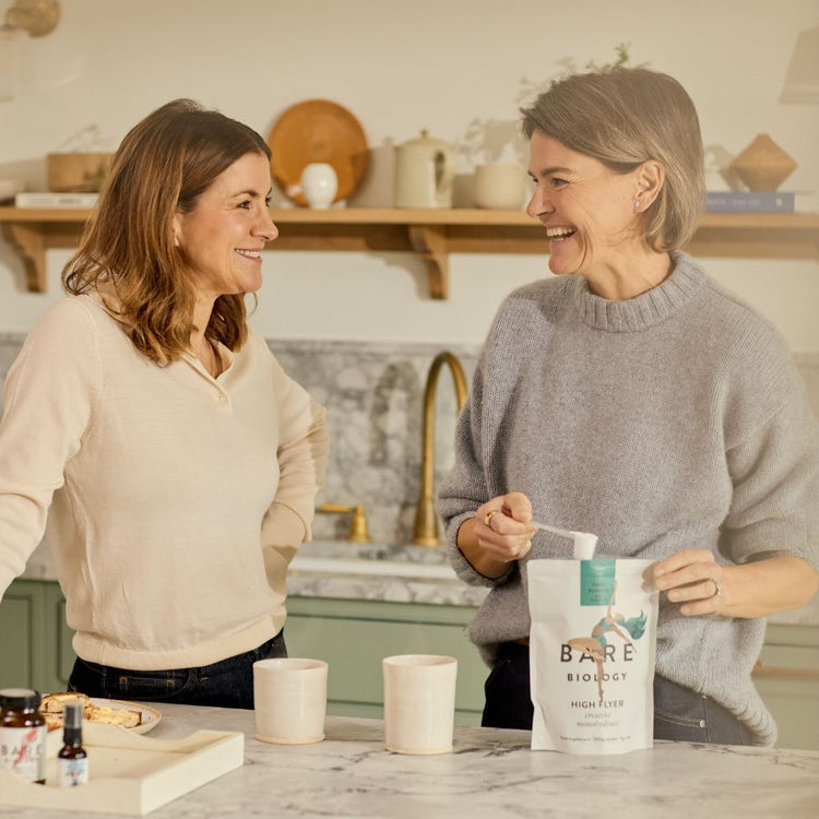 Two women looking at each other and smiling whilst one is scooping out Bare Biology Creatine Powder from the pouch in a beautiful kitchen setting.