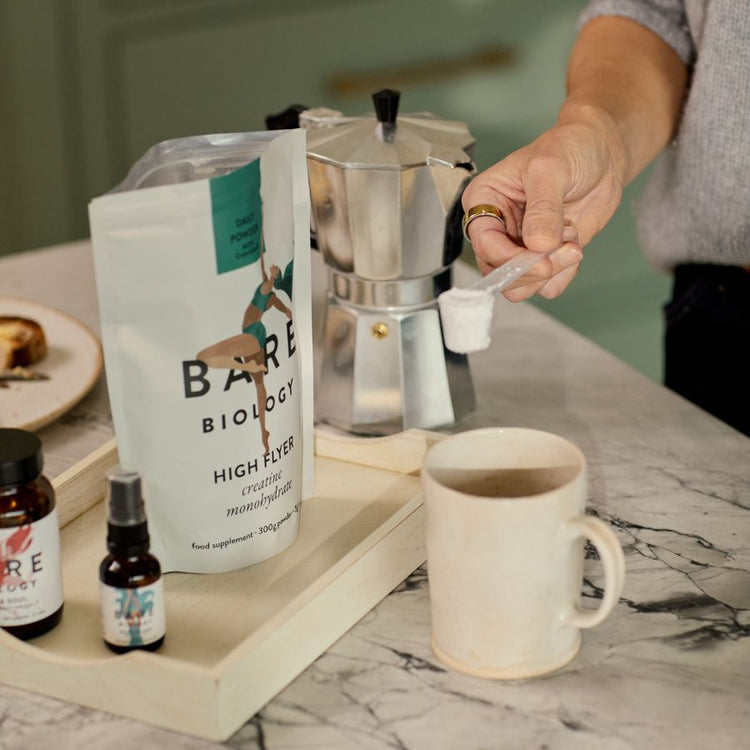Hand scooping Bare Biology creatine powder using a small plastic mug into a neutral coloured mug on a marbled kitchen counter.