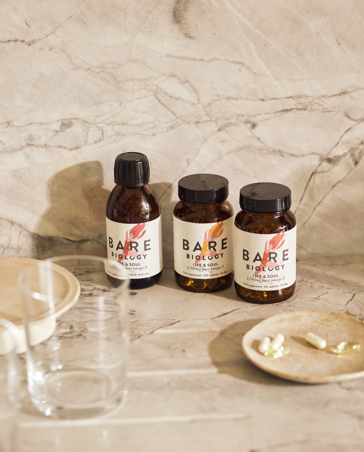 Three bottles of Bare Biology products on a marble surface with supplement capsules displayed on a small plate to the front on the right hand side