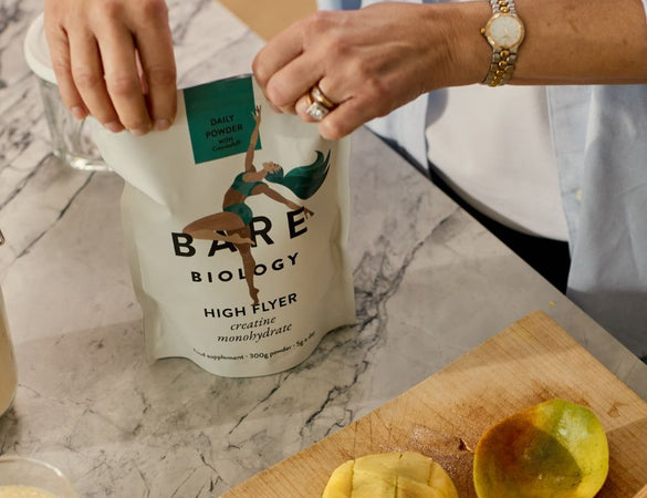 Person opening a Bare Biology High Flyer Creatine bag on a marble countertop with a cutting board and fruit.