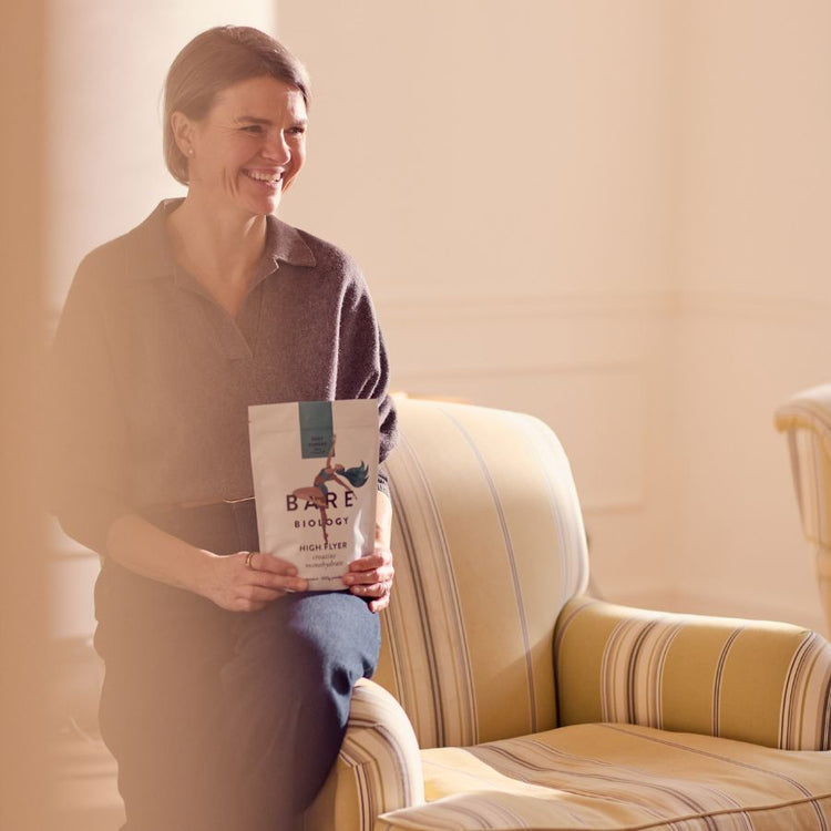 Mel the Founder and CEO of Bare Biology sitting on a striped fabric arm chair holding the new High Flyer Creatine Monohyrdrate product pouch in a warm-toned room.