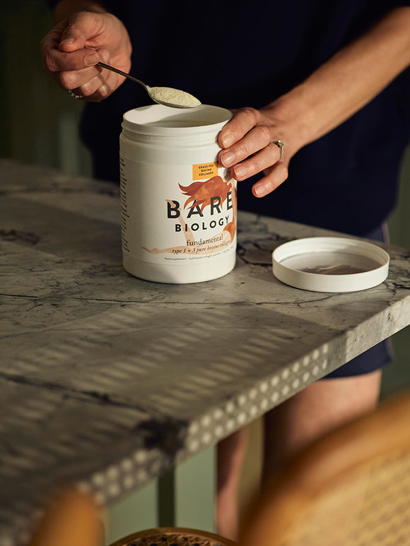 Woman scooping Bare Biology Fundamental Bovine Collagen powder out of tub with spoon at a counter top table
