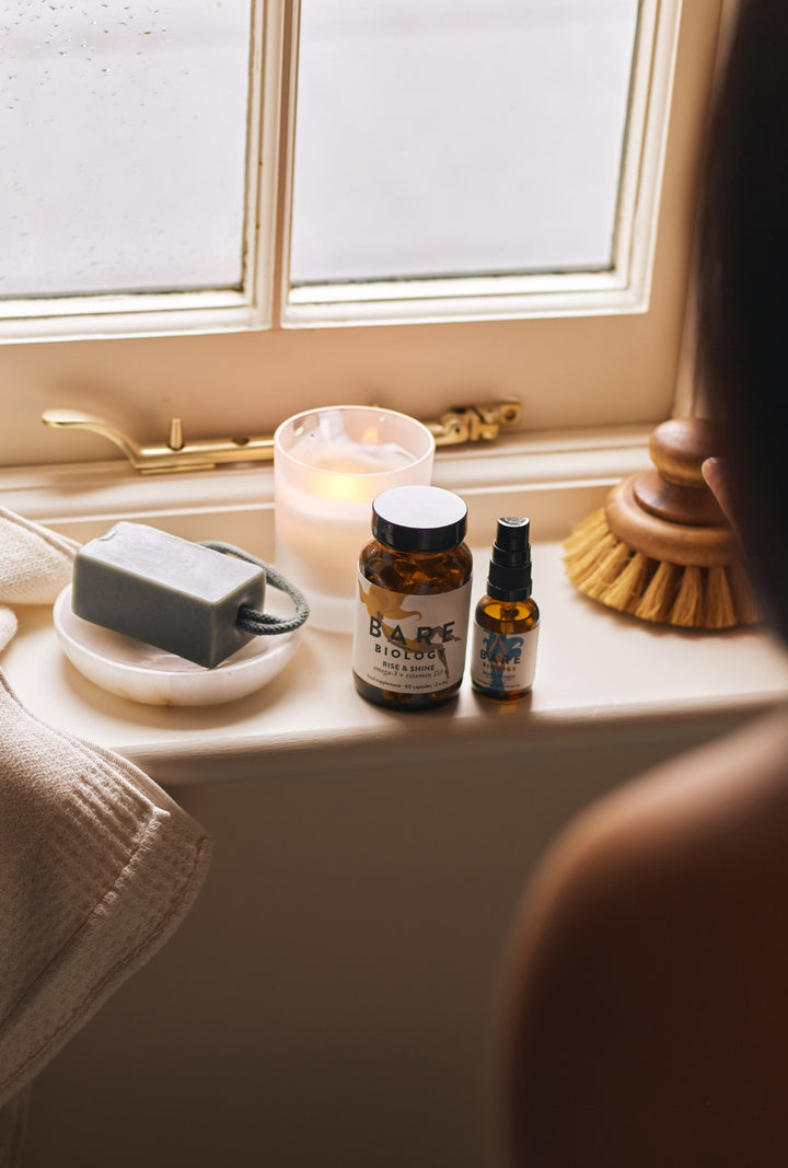 Bath products including a bottle and a glass bottle of Bare Biology fish oil + vitamin D capsules and Vitamin D spray on a windowsill with a candle and brush.