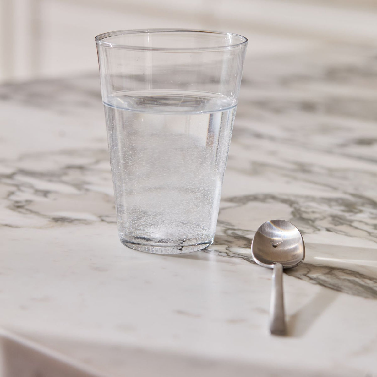 A clear glass tumbler filled with water and dissolved powder sits on a white marble surface next to a silver spoon.