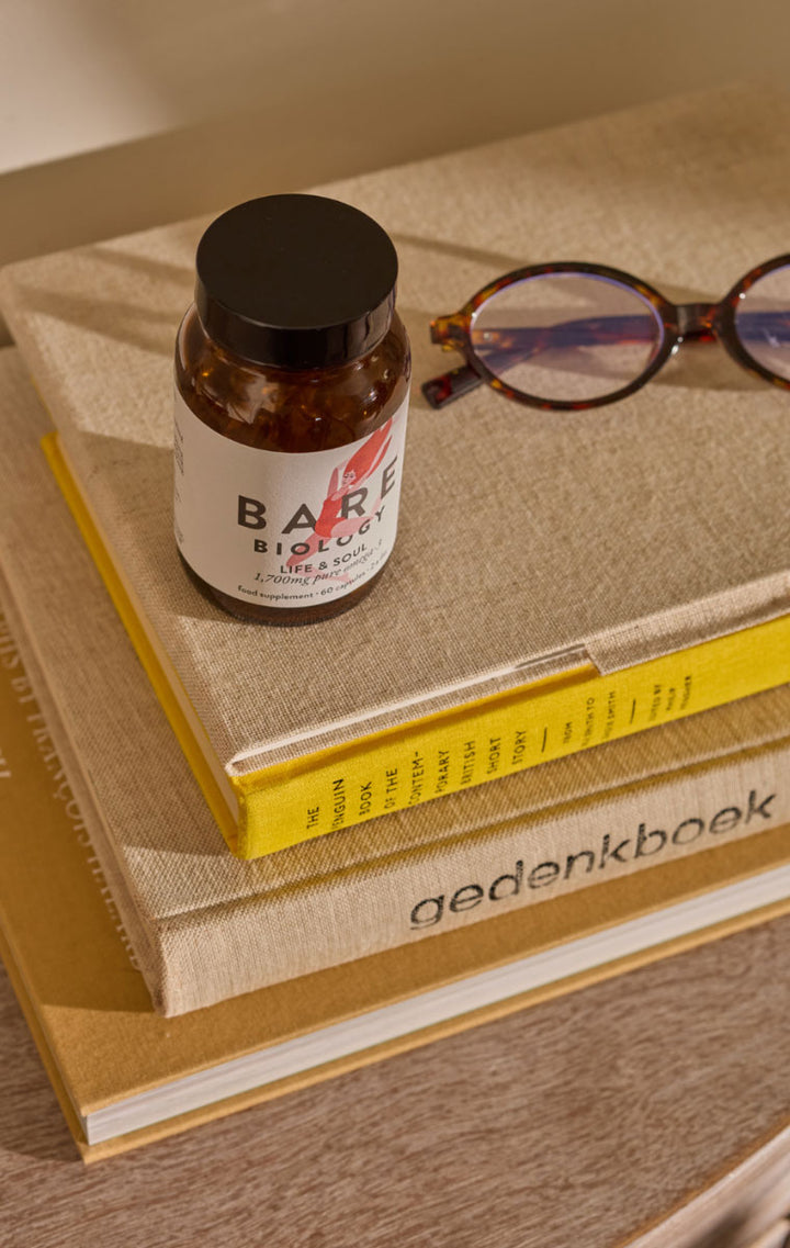 A small amber bottle labeled Bare Biology sits on top of stacked books, with tortoiseshell eyeglasses resting nearby. The scene is styled on a wooden surface with warm natural lighting.