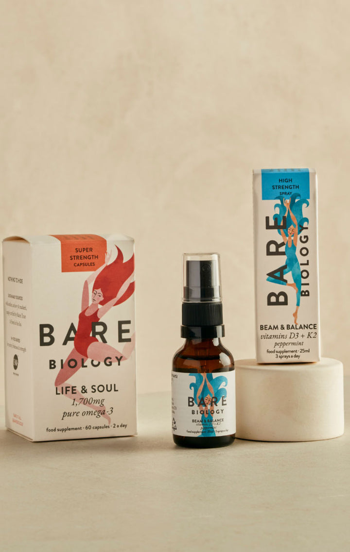 Bare Biology supplement products arranged on a beige surface against a matching beige background: a white box labeled Life & Soul, a small amber spray bottle with a black pump and white label, and a white box labeled Beam & Balance, with a white cylindrical container behind the bottle.