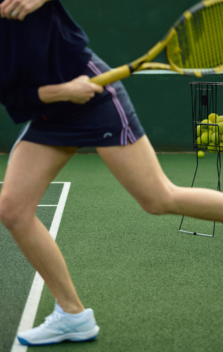 A close-up action shot of a tennis player on a green court, wearing a navy athletic skirt with pink stripes and light blue sneakers, holding a yellow tennis racket near a ball basket.