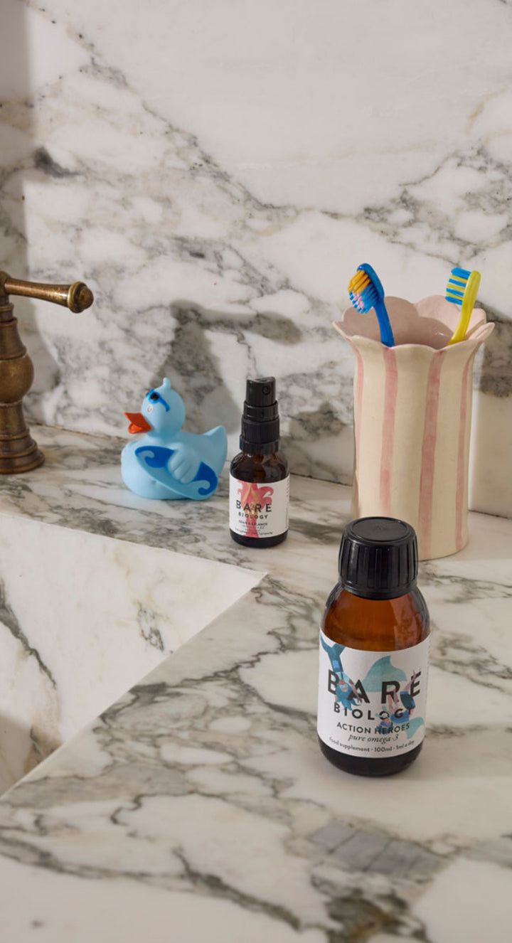 A small amber bottle labeled Bare Biology with a black cap sits on a white marble bathroom counter next to a brass faucet, a blue rubber duck, and a striped toothbrush holder with toothbrushes.