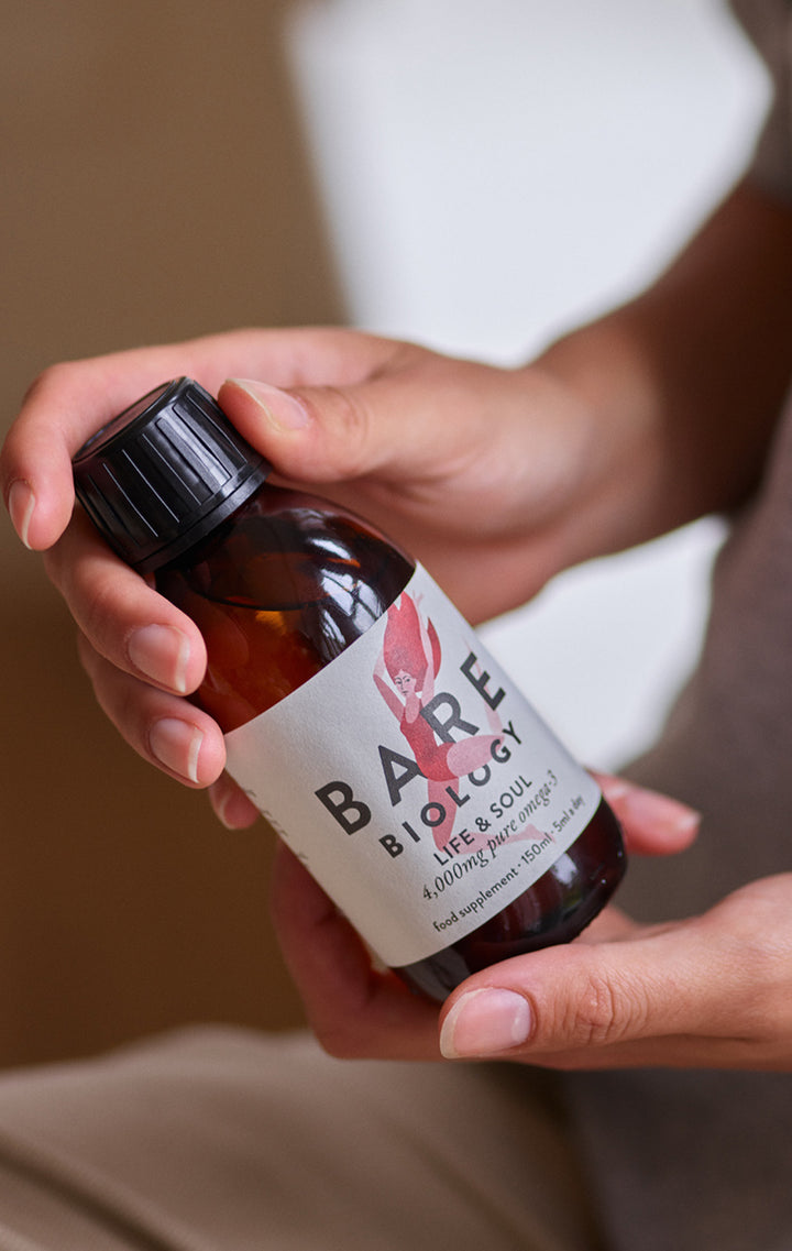 A person holding a small amber glass bottle labeled BARE BIOLOGY Life & Soul essential oil. The bottle has a black cap and a white label with a pink graphic, shown against a soft-focus indoor background.