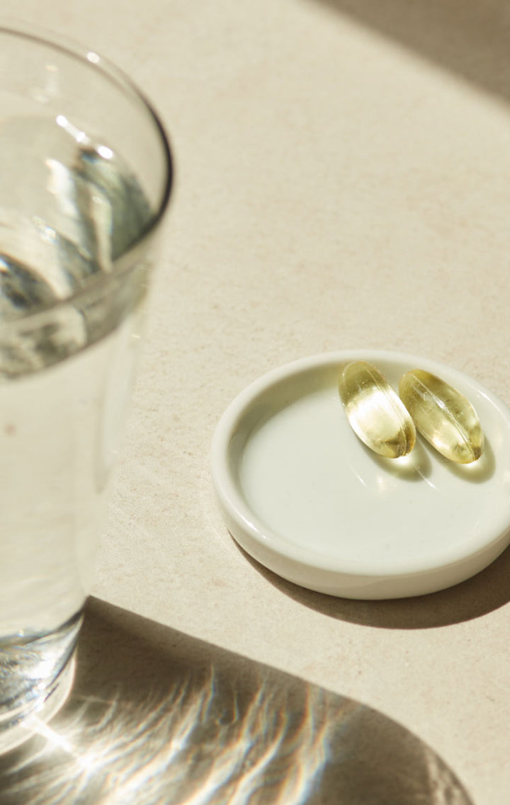 Two yellow softgel supplement capsules resting in a small white ceramic dish, next to a glass of water on a beige surface with warm natural sunlight and shadows.