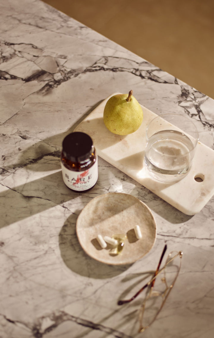 A small amber jar of Bare Biology Omega-3 product with a white label sits on a marble surface beside a pear, a glass of water, a shallow dish with capsules, and eyeglasses in a styled interior scene.