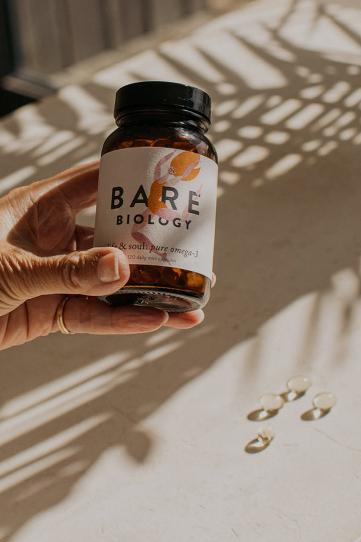 A woman's hand holding a jar of bare biology life & soul mini capsules. A shadow of a plant and 4 capsules in the background.