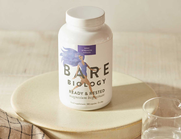 Bare Biology supplement bottle on a ceramic neutral coloured surface with a glass of water displayed to the right side of the product.